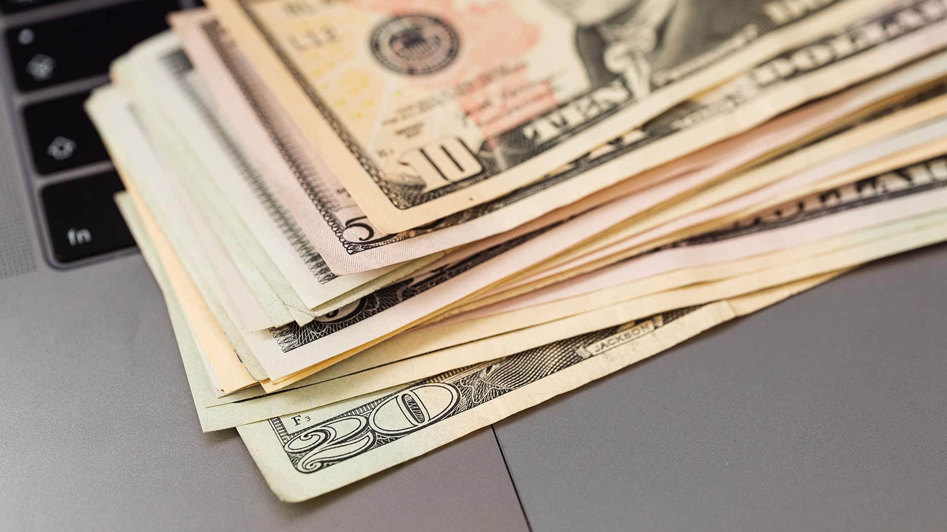 Stack of US dollar bills and financial documents on a laptop.